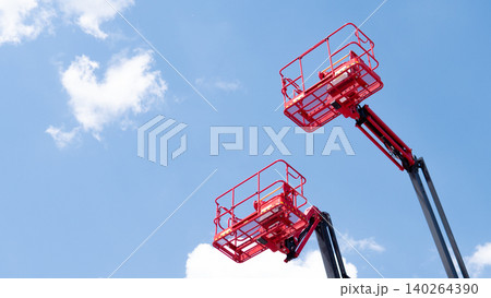Two red aerial work platforms raised high against a clear blue sky with soft white clouds, viewed from below in an industrial maintenance setting Two red aerial work platforms raised high against a clear blue sky with soft white clouds, viewed from below in an industrial maintenance setting 140264390