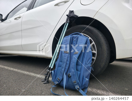 Blue backpack and trekking poles leaning against a white compact car parked on asphalt. High quality photo 140264457