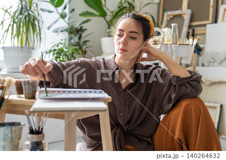Thoughtful woman painter painting picture in sketchbook with watercolor, enjoying creative process Thoughtful woman painter painting picture in sketchbook with watercolor, enjoying creative process 140264632