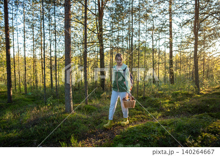 Woman walking in pine forest looks around, collecting wild fungi in wicker basket. Mushroom foraging 140264667