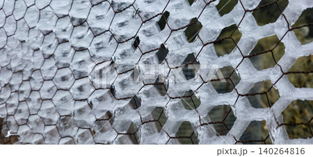 Ice formation on hexagonal wire mesh fence, showcasing intricate patterns and textures, with blurred natural background elements visible through the mesh. High quality photo Ice formation on hexagonal wire mesh fence, showcasing intricate patterns and textures, with blurred natural background elements visible through the mesh. High quality photo 140264816
