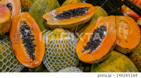 Freshly cut papaya fruit displayed among whole papayas in a grocery store, showcasing vibrant orange flesh and black seeds, with protective netting visible. High quality photo Freshly cut papaya fruit displayed among whole papayas in a grocery store, showcasing vibrant orange flesh and black seeds, with protective netting visible. High quality photo 140265184