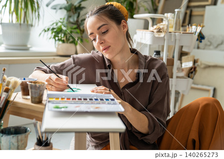 Cheerful inspired woman painter drawing sketches in notebook with aquarelle, fulfills ideas on paper Cheerful inspired woman painter drawing sketches in notebook with aquarelle, fulfills ideas on paper 140265273
