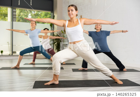 Active young European woman practicing Virabhadrasana 2 Warrior pose, indoor class with women, studio background 140265325