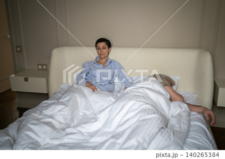 Senior woman with insomnia and sleep deprivation sitting in bed while husband sleeping and snoring. 140265384