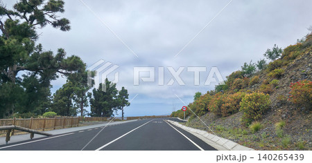 Scenic view of a winding road bordered by lush greenery and trees, leading towards the horizon under a cloudy sky in a mountainous region of island La Palma. High quality photo 140265439