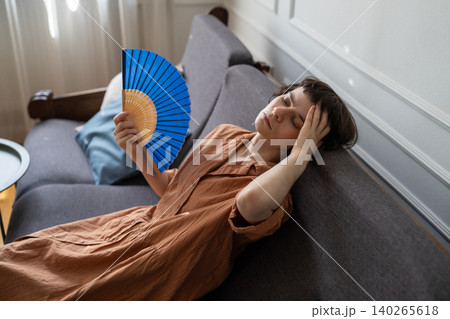Stressed woman suffering from summer heat and migraine at home, lying on sofa waving paper fan. Stressed woman suffering from summer heat and migraine at home, lying on sofa waving paper fan. 140265618