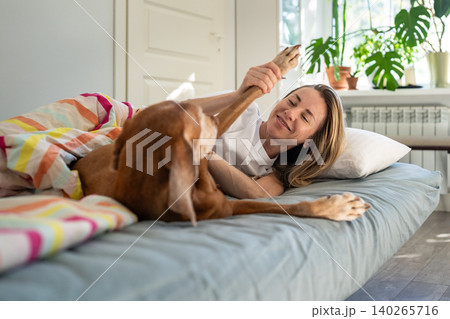Laughing happy woman lying in bed with Vizsla dog, fooling playing around with pet on weekend. Laughing happy woman lying in bed with Vizsla dog, fooling playing around with pet on weekend. 140265716