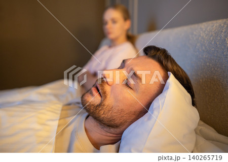 Man sleeping in bed and snoring loud while frustrated wife sitting beside with exhausted expression. Man sleeping in bed and snoring loud while frustrated wife sitting beside with exhausted expression. 140265719