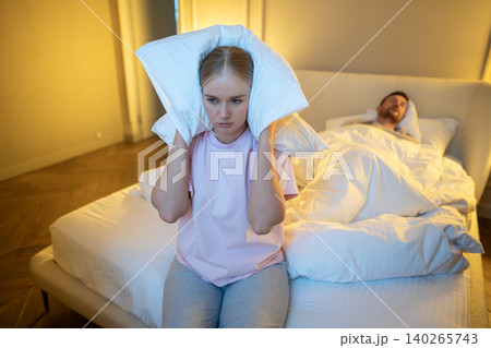 Irritated wife sitting on bed covering ears with pillow to block snore sound while man sleeping. Irritated wife sitting on bed covering ears with pillow to block snore sound while man sleeping. 140265743