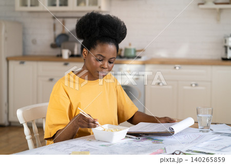 African girl student study exam has lunch reading documents at kitchen table with blueprints drafts African girl student study exam has lunch reading documents at kitchen table with blueprints drafts 140265859