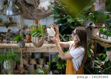 Woman gardener in overalls watering potted houseplant in greenhouse surrounded by plants and pots 140265868