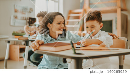 Smiling Hispanic adult teacher helping young boy write on paper in a sunny school classroom 140265929