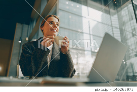 Smiling young adult Caucasian woman drinking coffee near laptop in a modern city cafe Smiling young adult Caucasian woman drinking coffee near laptop in a modern city cafe 140265973