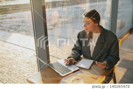 Caucasian young adult woman working on laptop inside bright sunny cafe next to large window Caucasian young adult woman working on laptop inside bright sunny cafe next to large window 140265978