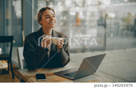 Caucasian young woman smiling while holding coffee cup next to laptop in a cafe Caucasian young woman smiling while holding coffee cup next to laptop in a cafe 140265979