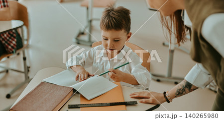 Caucasian young boy writing in notebook with teacher helping in bright classroom Caucasian young boy writing in notebook with teacher helping in bright classroom 140265980