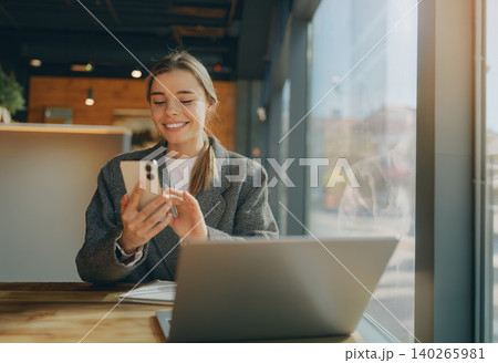 Caucasian young adult woman smiling while using smartphone inside modern cafe next to laptop Caucasian young adult woman smiling while using smartphone inside modern cafe next to laptop 140265981
