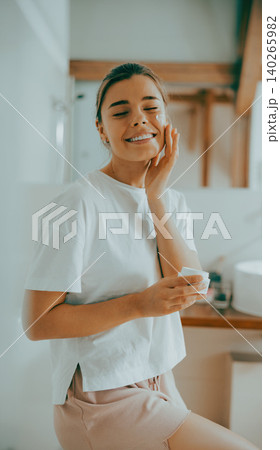 Happy young Caucasian woman in her twenties applying face moisturizer cream in bright bathroom 140265982
