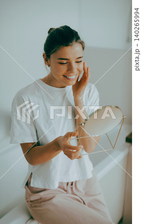 Happy young Caucasian woman looking in hand mirror touching her cheek in a bright bathroom Happy young Caucasian woman looking in hand mirror touching her cheek in a bright bathroom 140265994