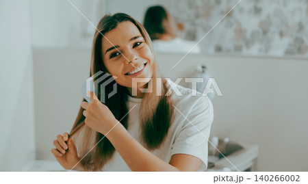 Smiling young adult Caucasian woman brushing her long brown hair in a bright modern bathroom Smiling young adult Caucasian woman brushing her long brown hair in a bright modern bathroom 140266002