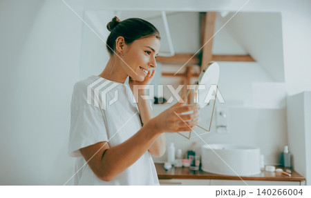 Happy young adult Caucasian woman looking at her face in a hand mirror in a bright bathroom Happy young adult Caucasian woman looking at her face in a hand mirror in a bright bathroom 140266004