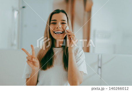 Happy young adult Caucasian woman cleanses face and makes peace sign in bright bathroom Happy young adult Caucasian woman cleanses face and makes peace sign in bright bathroom 140266012