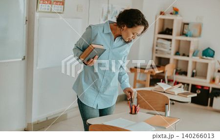 Middle aged Latina teacher holding book and arranging pens on desk in classroom Middle aged Latina teacher holding book and arranging pens on desk in classroom 140266013