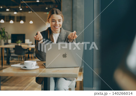 Happy young Caucasian woman gestures during video call on laptop inside cozy cafe 140266014