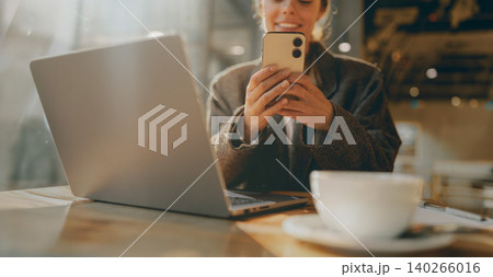 Caucasian young adult woman using her smartphone next to a laptop inside a warm cafe 140266016