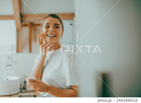 Happy young Caucasian woman in her twenties applying skin cream in a bright bathroom Happy young Caucasian woman in her twenties applying skin cream in a bright bathroom 140266018