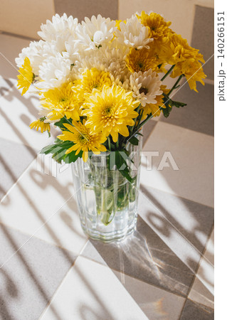 Bouquet of fresh white and yellow chrysanthemum flowers in a glass vase standing on a tiled balcony floor with sharp contrast shadows from a railing in sunlight Bouquet of fresh white and yellow chrysanthemum flowers in a glass vase standing on a tiled balcony floor with sharp contrast shadows from a railing in sunlight 140266151