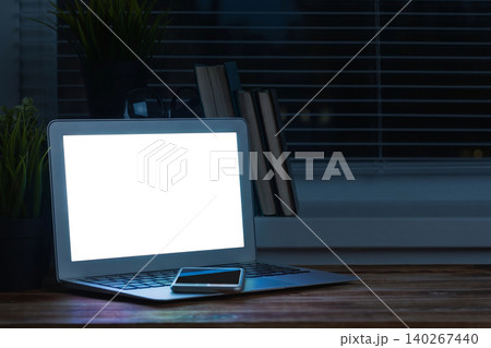 Workstation setup with laptop and smartphone at night near the window and bookshelf Workstation setup with laptop and smartphone at night near the window and bookshelf 140267440