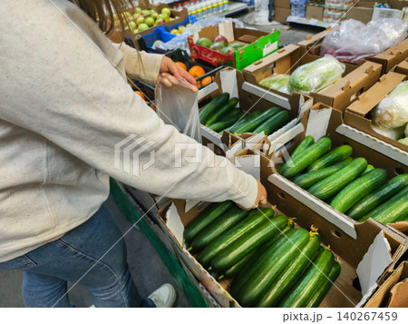Woman's hand reaching into a cardboard crate of fresh green cucumbers at a supermarket produce aisle, selecting healthy ingredients for cooking and a plant-based diet. Woman's hand reaching into a cardboard crate of fresh green cucumbers at a supermarket produce aisle, selecting healthy ingredients for cooking and a plant-based diet. 140267459