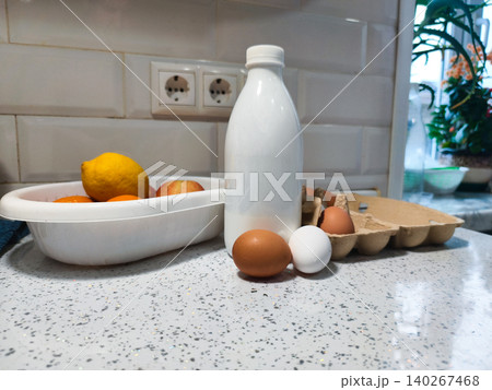 Daily fresh milk bottle, brown and white eggs in an open carton, and various fresh fruits stored in a plastic bowl on a speckled white kitchen countertop, creating a rustic still life. Daily fresh milk bottle, brown and white eggs in an open carton, and various fresh fruits stored in a plastic bowl on a speckled white kitchen countertop, creating a rustic still life. 140267468