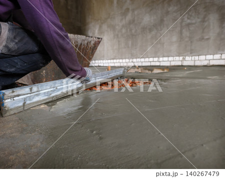 Construction worker in protective gloves leveling freshly poured concrete screed on a building site, ensuring a smooth and even surface for the new floor installation. Construction worker in protective gloves leveling freshly poured concrete screed on a building site, ensuring a smooth and even surface for the new floor installation. 140267479