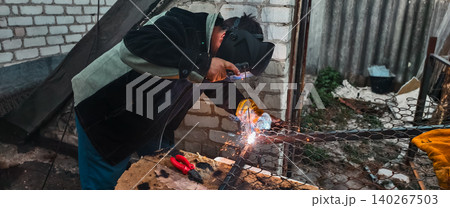Person wearing protective gear, including a welding mask and gloves, using an arc welder to join metal parts, generating bright sparks during the repair process outdoors. 140267503