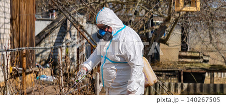 Person wearing a protective hazmat suit, respirator mask, and gloves spraying chemicals on dormant garden plants, protecting against pests and diseases in a rural backyard setting. Person wearing a protective hazmat suit, respirator mask, and gloves spraying chemicals on dormant garden plants, protecting against pests and diseases in a rural backyard setting. 140267505