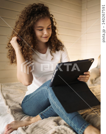 Young woman engaged with tablet while sitting on bed in cozy room during daytime Young woman engaged with tablet while sitting on bed in cozy room during daytime 140267591