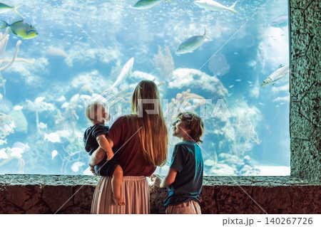 Family sits together by a bright marine tank, showing ocean education, travel with kids, sibling curiosity and underwater wildlife concept Family sits together by a bright marine tank, showing ocean education, travel with kids, sibling curiosity and underwater wildlife concept 140267726