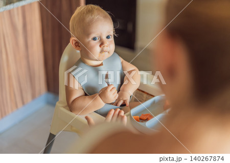 Infant practices self feeding with vegetables in a kitchen, showing baby led weaning, early nutrition and family care concept Infant practices self feeding with vegetables in a kitchen, showing baby led weaning, early nutrition and family care concept 140267874