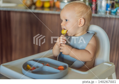 Infant practices self feeding with vegetables in a kitchen, showing baby led weaning, early nutrition and family care concept Infant practices self feeding with vegetables in a kitchen, showing baby led weaning, early nutrition and family care concept 140267881