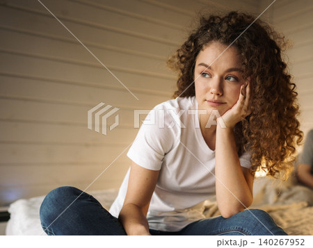 Young woman sitting on bed with thoughtful expression in cozy room during daytime 140267952