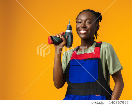 Young person holds a drill and smiles against a bright orange background in a workshop setting Young person holds a drill and smiles against a bright orange background in a workshop setting 140267995