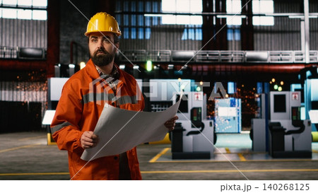 Engineer walking along facility reviewing manufacturing blueprints and technical plans, heavy industry warehouse. Production hall features machinery instructions, layout drawings. Engineer walking along facility reviewing manufacturing blueprints and technical plans, heavy industry warehouse. Production hall features machinery instructions, layout drawings. 140268125