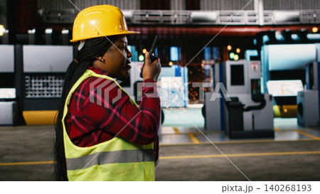 Black woman engineer speaking into portable walkie-talkie device, walking through warehouse and monitoring heavy equipment. Large-scale industrial facility highlights production line. 140268193
