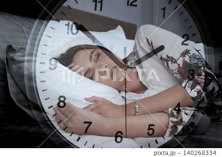 Woman sleeps on bed while clock shows time on a busy evening in a room 140268334