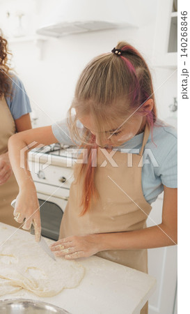 Girl cuts dough while cooking with adult in a bright kitchen during the day Girl cuts dough while cooking with adult in a bright kitchen during the day 140268846
