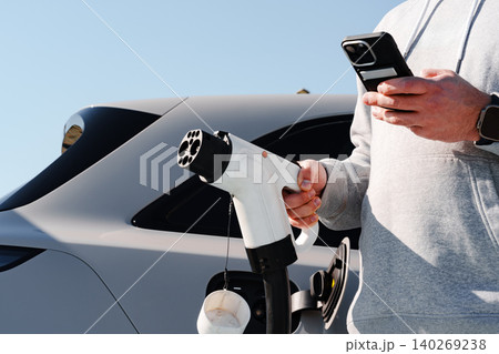 Charging an electric car while checking a smartphone during the day Charging an electric car while checking a smartphone during the day 140269238
