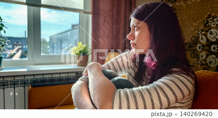 Teen girl with purple hair sitting on a couch, hugging her knees while gazing out the window in a cozy living room with plants and warm lighting Teen girl with purple hair sitting on a couch, hugging her knees while gazing out the window in a cozy living room with plants and warm lighting 140269420